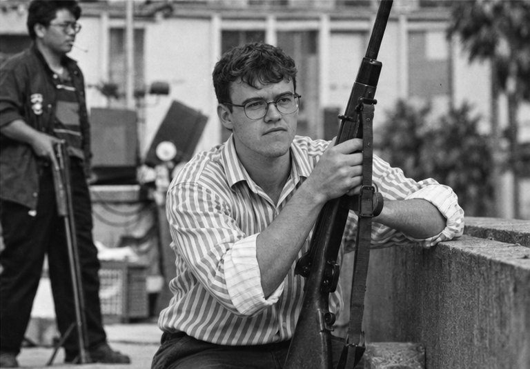 joakim .h in a striped shirt, crouched behind a low wall holding an old rifle